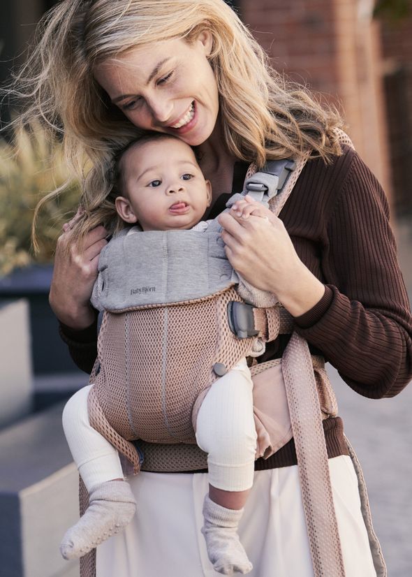 A smiling woman holds a baby in a carrier, with the baby sticking out their tongue. They are outdoors with blurred background elements.