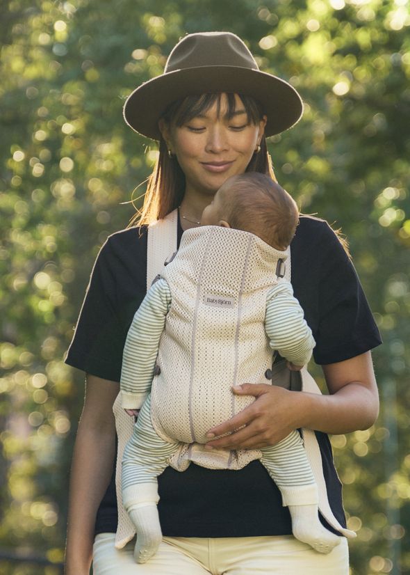 A woman wearing a hat and black shirt carries a baby in a front-facing carrier while standing outdoors with a blurred green background.