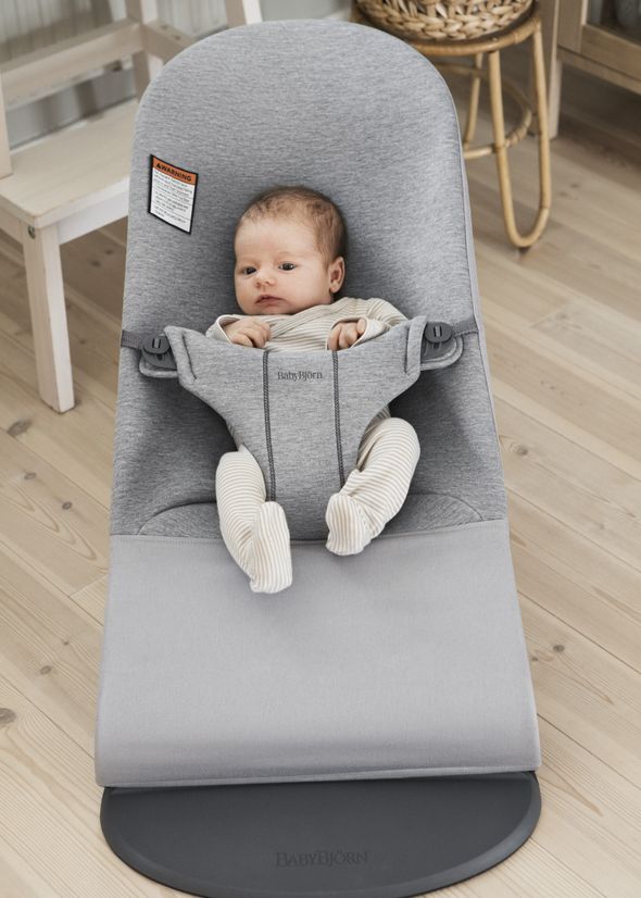 A baby in a gray baby bouncer on a wooden floor, wearing a striped outfit, looking up with a calm expression.