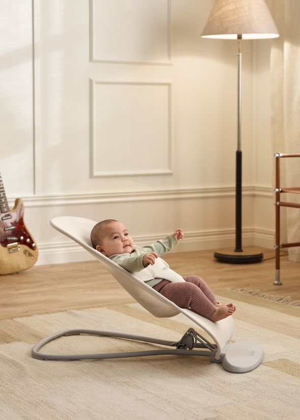 A baby relaxing in a cushioned baby bouncer on a rug, with a guitar in the background and a floor lamp nearby.