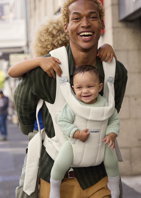 Man smiling, walking on city street with a baby in a front carrier and a child playfully hugging him from behind.