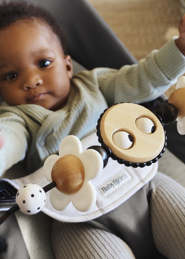 Baby in a BabyBjörn carrier reaching toward a wooden and white flower toy on a play bar across the front.