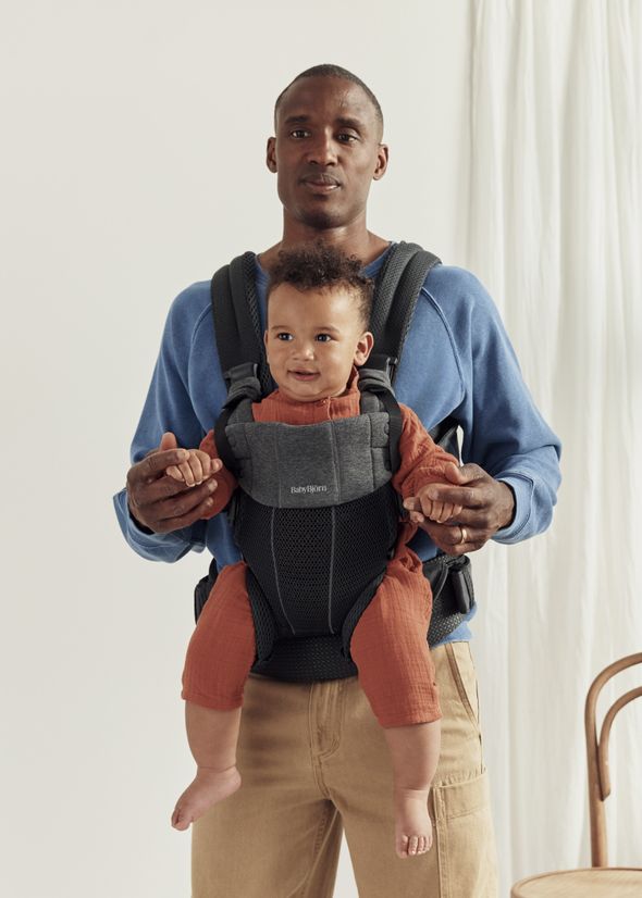 A man wearing a blue shirt holds a baby in a gray baby carrier. The baby, in a rust-colored outfit, is smiling and looking forward.