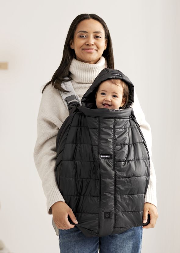 A woman wearing a beige turtleneck carries a smiling baby in a black baby carrier with a hood, standing indoors.