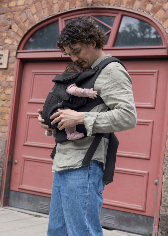 A person stands in front of a red door, wearing a baby carrier holding an infant.