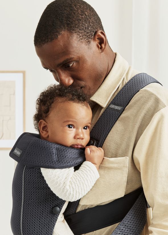 A man lovingly looks at a baby in a black carrier. The baby is held close to his chest, wearing a white sweater in a cozy, light-filled room.