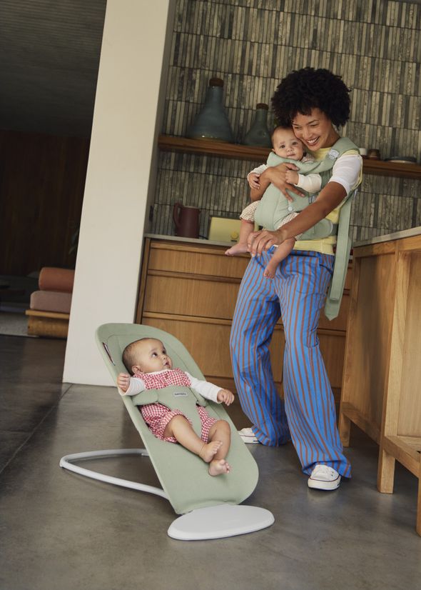 A person holding a baby in a carrier smiles at another baby sitting in a green baby bouncer. They're in a modern kitchen.