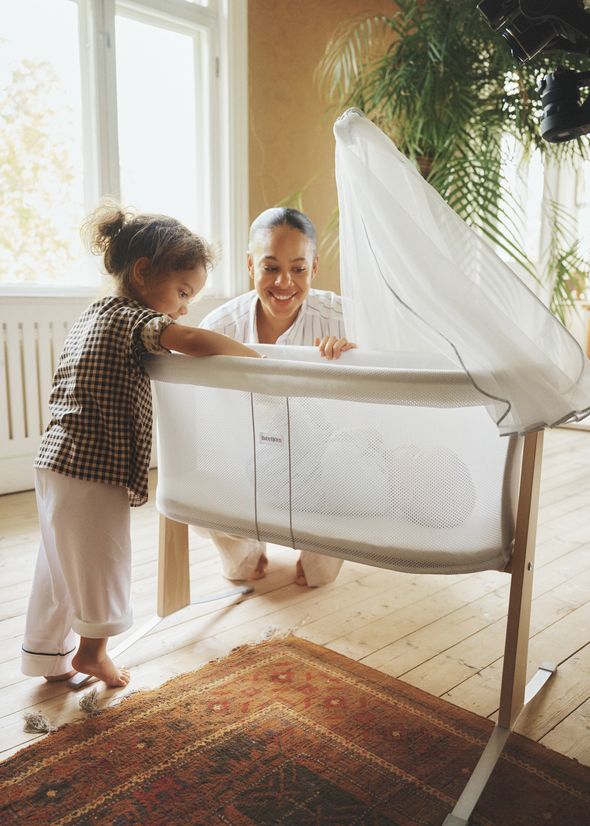 A smiling woman and a young child look at a baby sleeping in a white bassinet in a sunlit room with plants and wooden flooring.