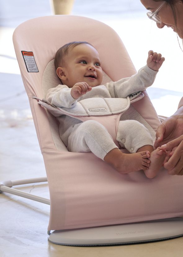 A smiling baby in a pink bouncer, wearing a light gray outfit, interacts with an adult holding their feet.