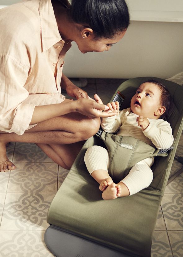 Woman crouching beside a baby in a bouncer, holding the baby's hand and smiling. The baby looks up at her with curiosity.