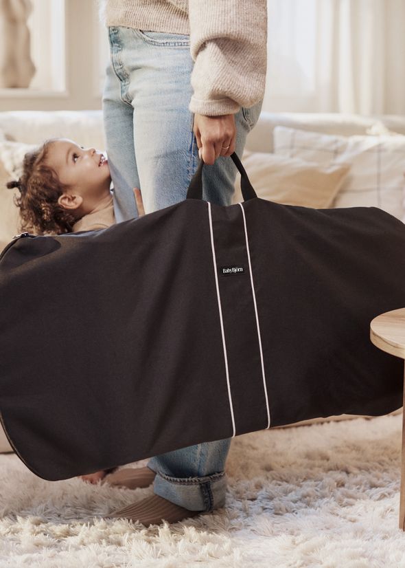 Child hugging an adult's leg in jeans, while the adult holds a black BabyBjörn bouncer bag, in a cozy room with soft lighting.