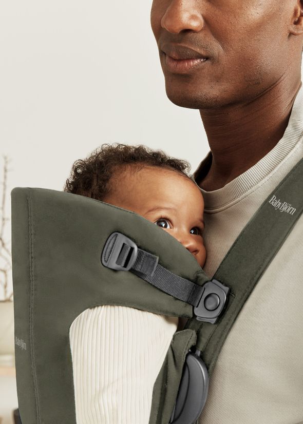 A baby in a beige outfit is securely held in a dark green baby carrier worn by an adult, both facing forward in a softly lit room.