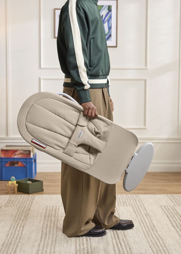 Person in a green jacket holding a beige baby bouncer sideways in a cozy room with framed art and toys in the background.
