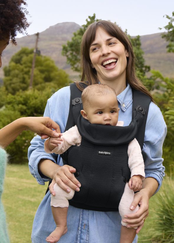 A woman smiling while carrying a baby in a black carrier outdoors, with another person reaching out to the baby.