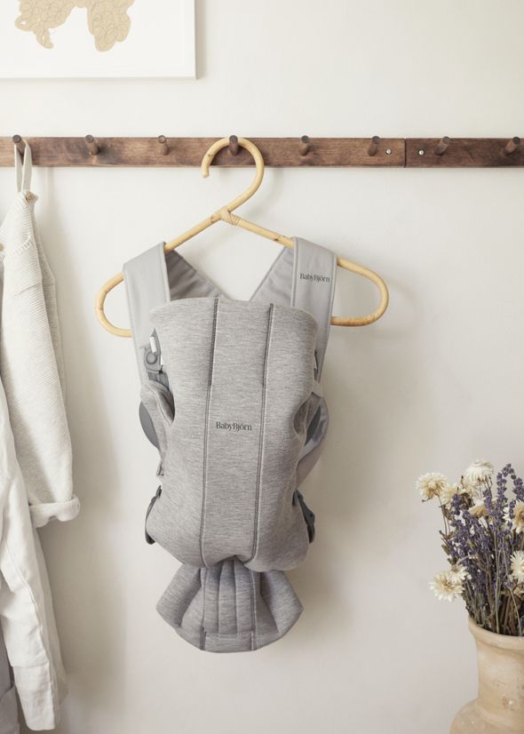 Gray baby carrier hanging on a wooden peg, with a beige sweater beside it and dried flowers in a vase to the right.