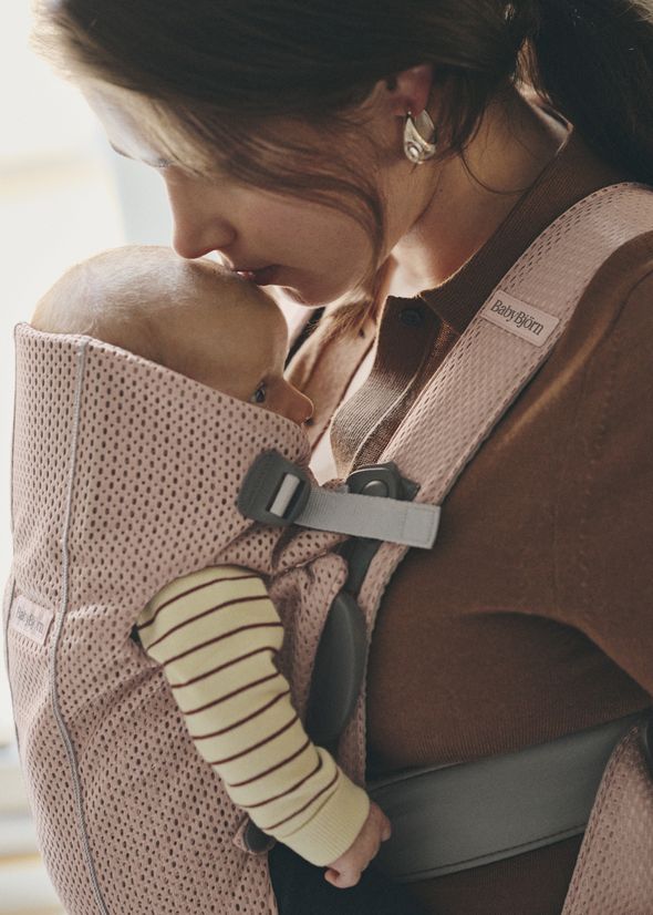 A woman kisses a baby's head while carrying them in a pink BabyBjörn carrier. The baby wears a striped outfit.