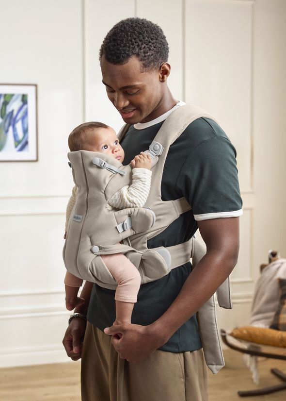 A man smiles at a baby carried in a beige baby carrier. The baby looks up at him in a cozy room with a framed artwork on the wall.