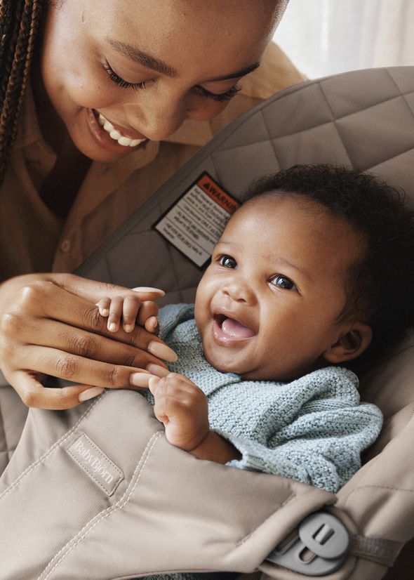 Smiling woman gently holds the hand of a laughing baby in a car seat, both appearing joyful and content.