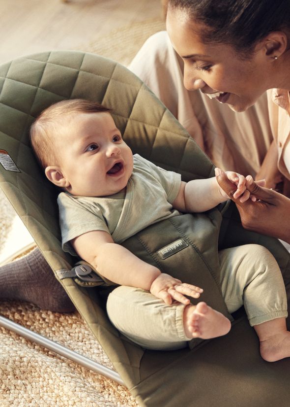 A smiling baby in a green bouncer looks up at a woman who is gently holding the baby's hand. They are both indoors on a light-colored rug.