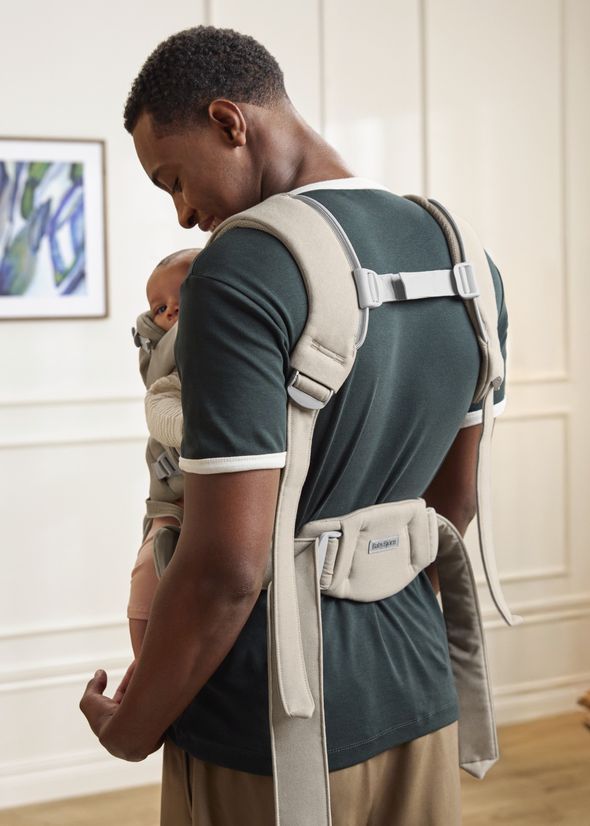 Man wearing a green shirt and khaki pants carries a baby in a beige baby carrier in a bright room with a painting on the wall.