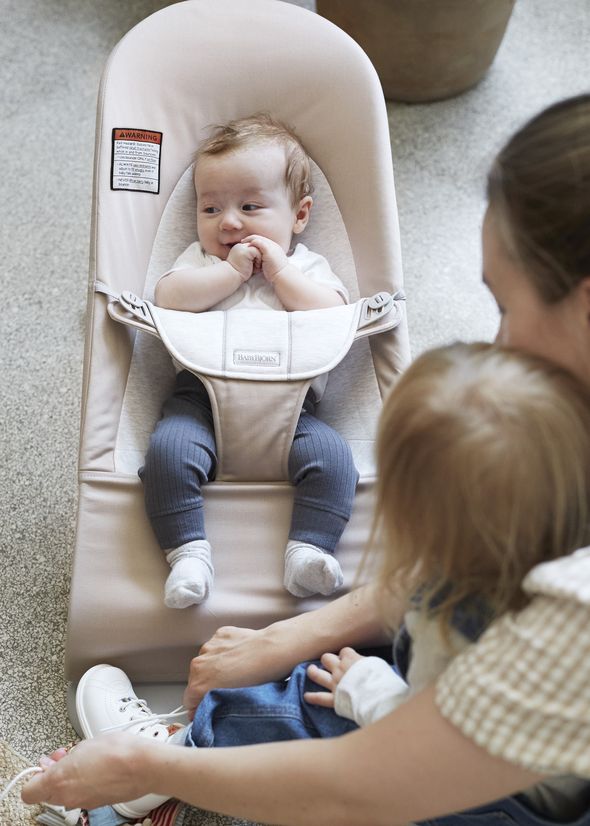 A baby sits in a beige bouncer, looking up, while a woman with a toddler adjusts a shoe. The setting is indoors with a soft, neutral background.
