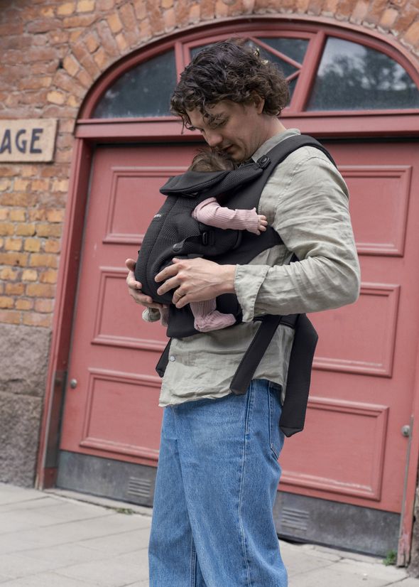 A man stands in front of a red brick building, gently holding a baby in a black carrier strapped to his chest.