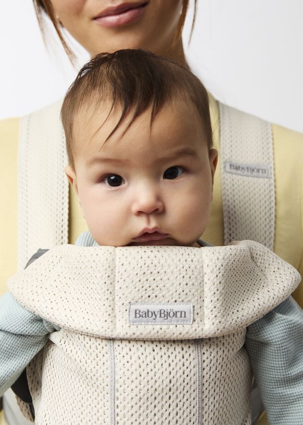 A baby in a cream-colored BabyBjörn carrier, facing forward. The carrier features a mesh design. An adult holds them from behind.