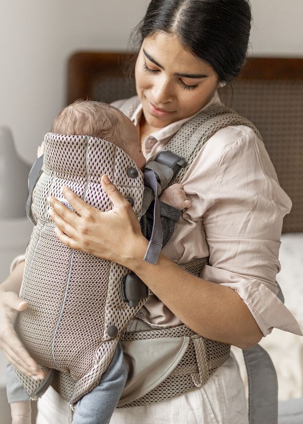 A woman gently holds a baby in a gray mesh baby carrier, smiling softly while standing indoors.