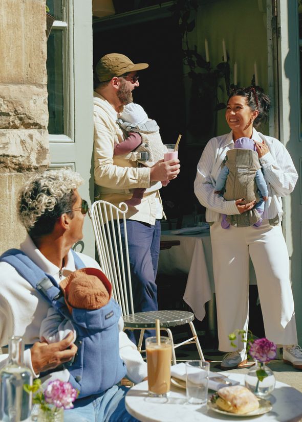 Three adults with baby carriers chat outside a café, enjoying drinks. A table with iced coffee and flowers is in the foreground.