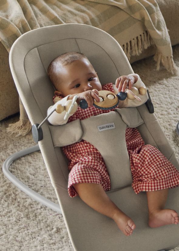 A baby in a red checkered outfit sits in a beige baby bouncer begin, playing with a wooden toy bar Googly eyes. The bouncer is on a soft carpet.
