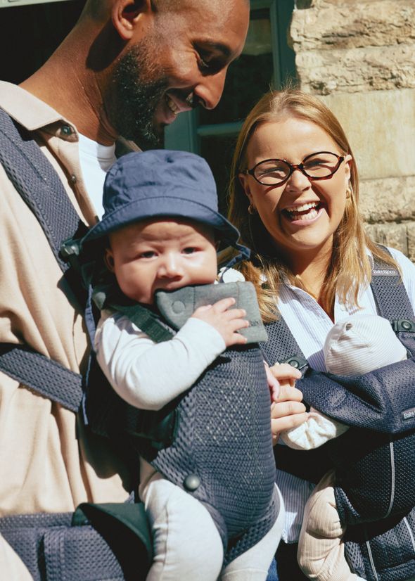 A smiling couple stands outside with two babies in front carriers. The woman wears glasses, and one baby is wearing a blue hat.