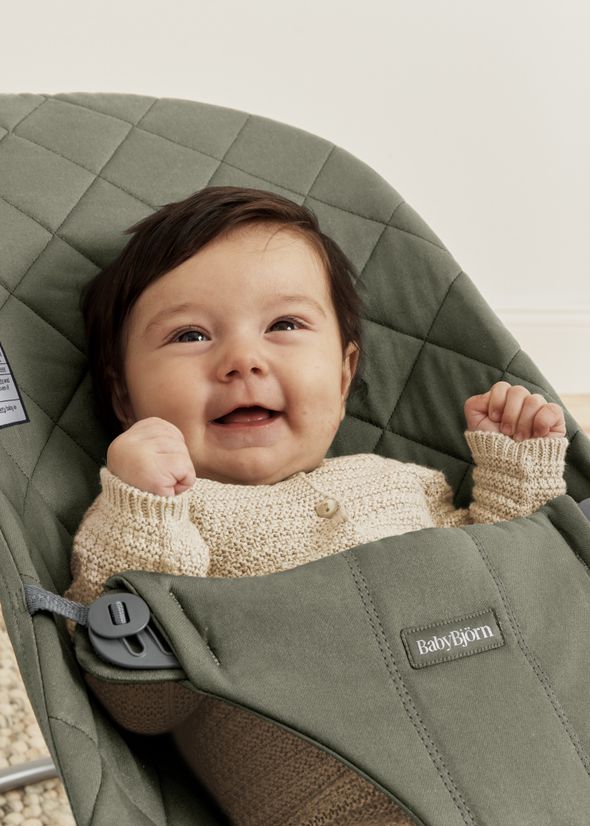 A smiling baby in a beige sweater sits in a green quilted baby bouncer, with fists raised in excitement.