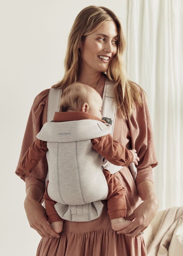 Woman smiling while carrying a sleeping baby in a gray BabyBjörn carrier, both wearing matching brown outfits.