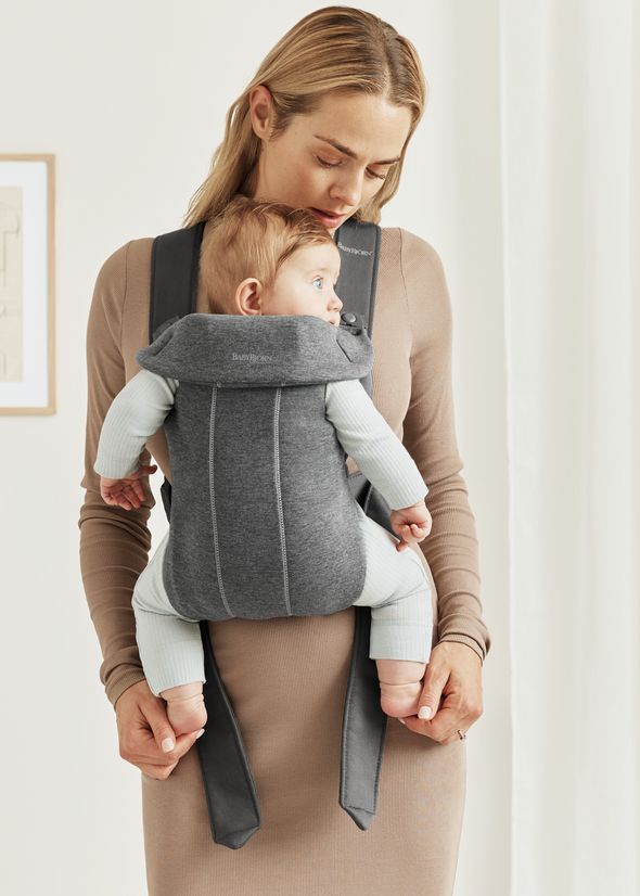 A woman wearing a beige dress holds a baby in a gray BabyBjörn carrier, looking down affectionately.