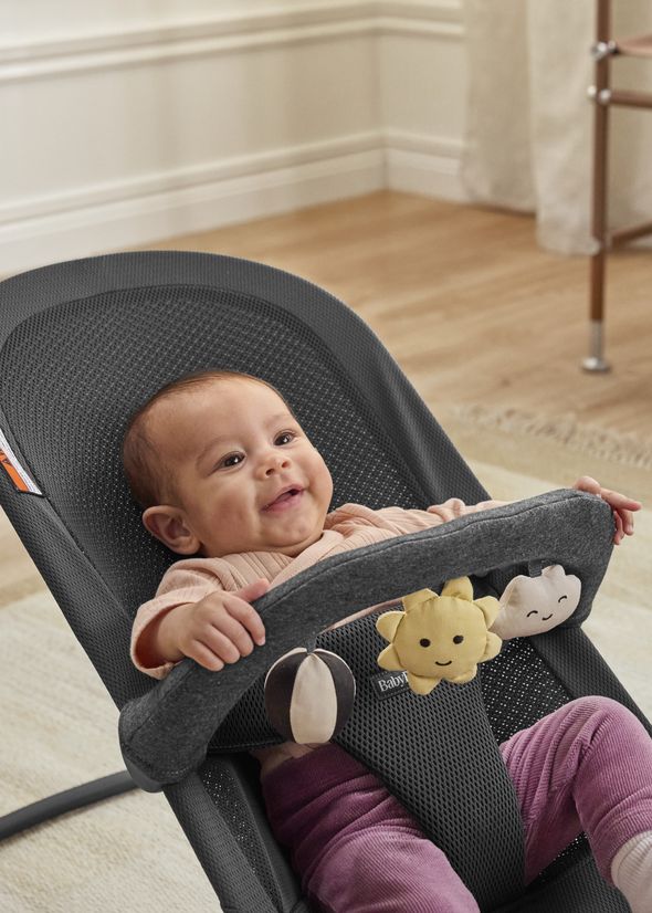 A smiling baby in a bouncer seat with hanging toys shaped like a sun, cloud, and ball, set in a cozy, light-colored room.