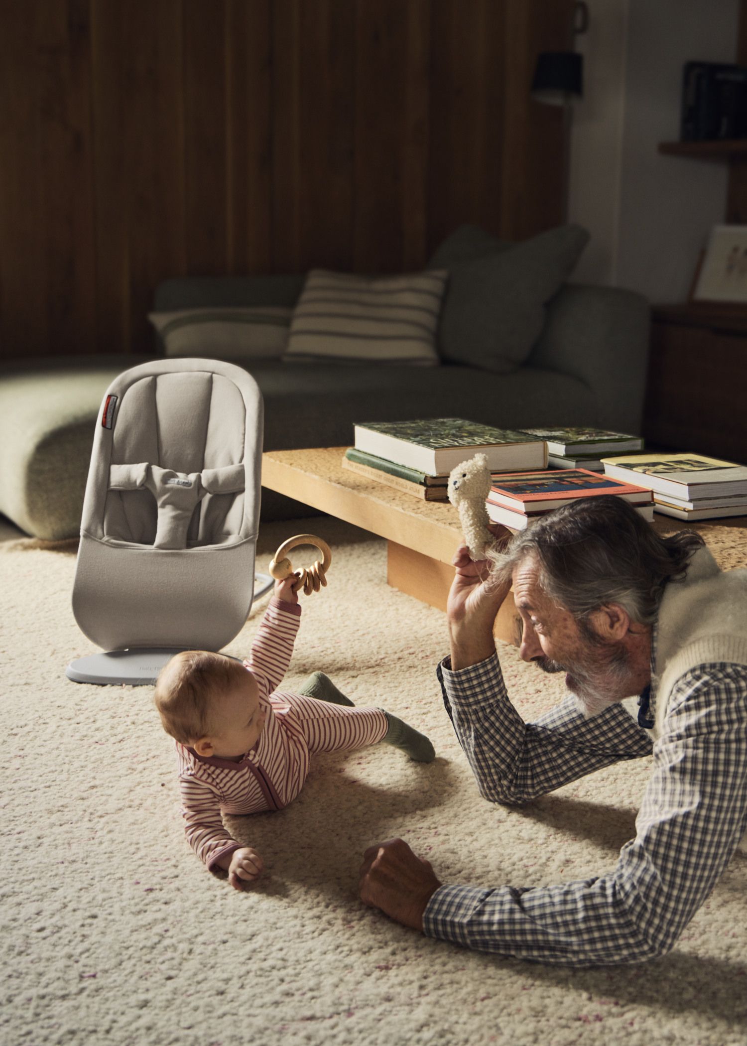 A baby in striped pajamas plays on the carpet with an elderly man holding a stuffed toy, next to a baby chair and a coffee table with books.