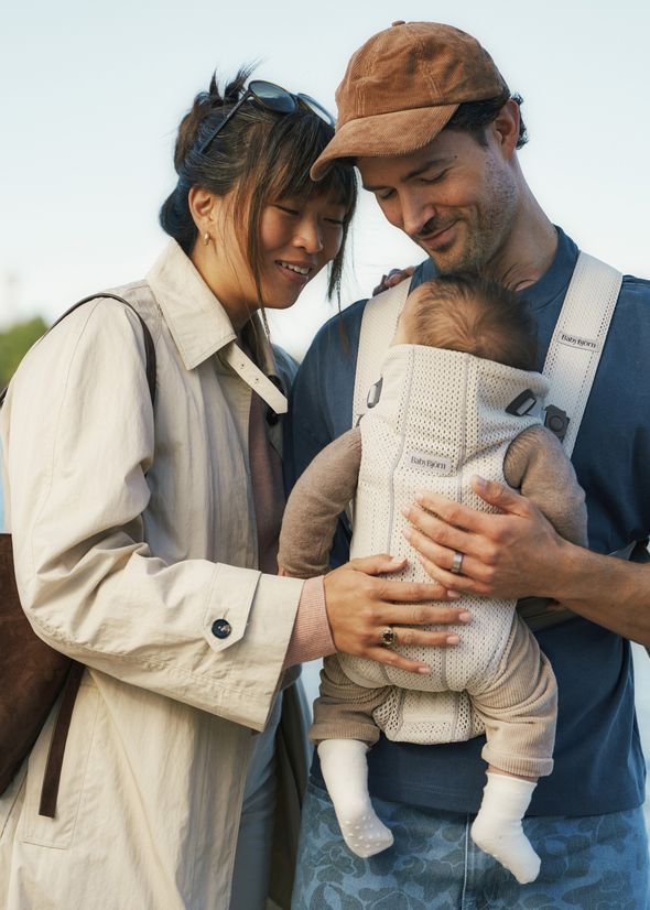 A couple smiles at their baby in a carrier. The mother wears a light coat and the father a cap, looking lovingly at the infant dressed in beige.