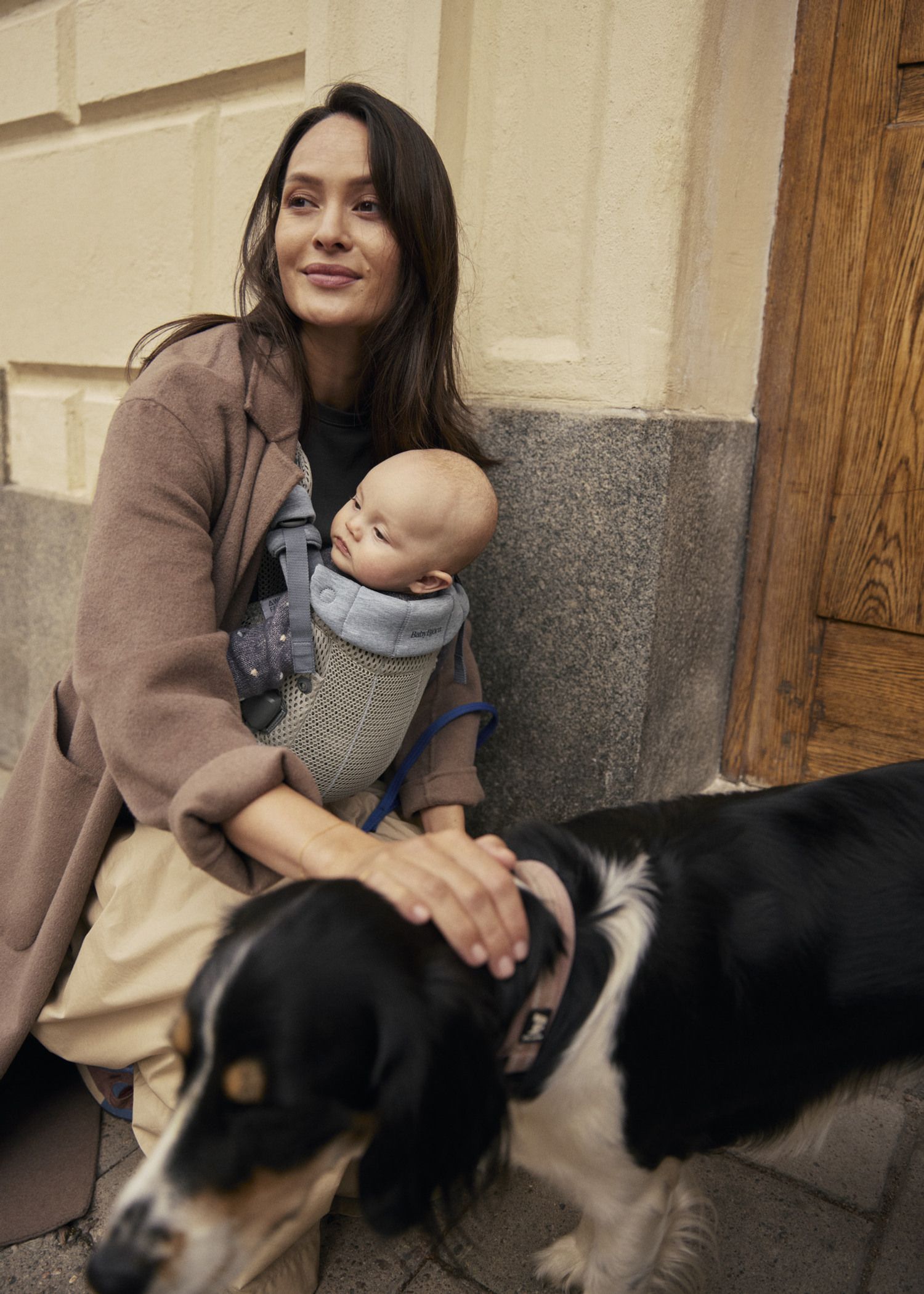 Woman sitting with a baby in a carrier, gently petting a black and white dog, against a building wall.