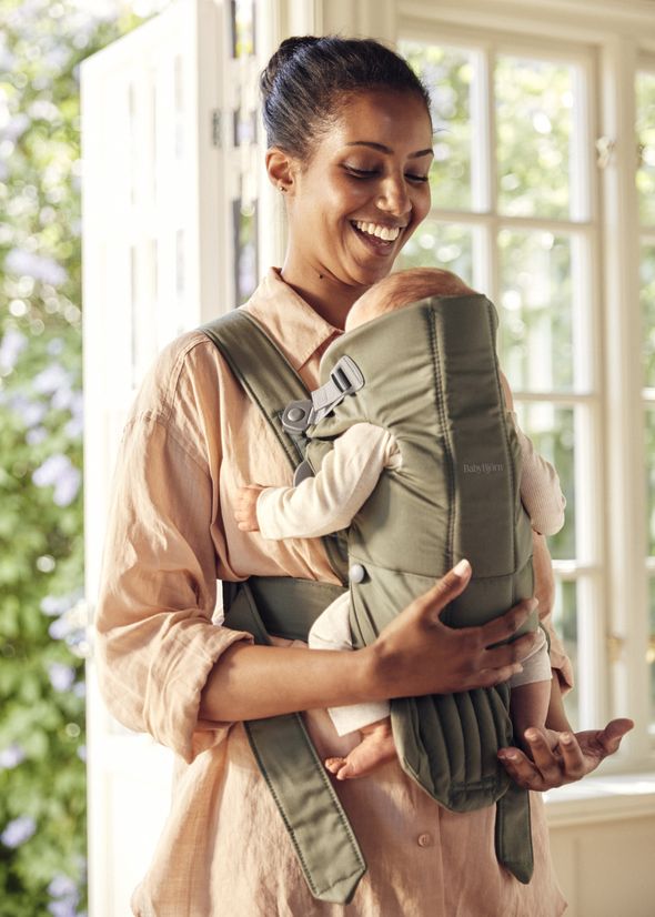 A smiling woman holds a baby in a green baby carrier inside a sunlit room with open windows and lush greenery outside.