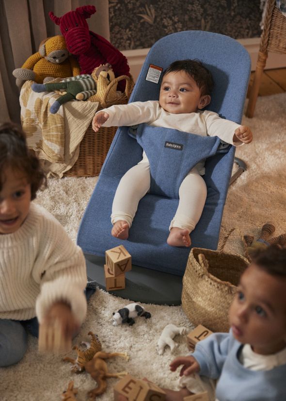 Three children playing on a soft carpet. One baby in a blue bouncer, and two toddlers playing with wooden blocks and toy animals.