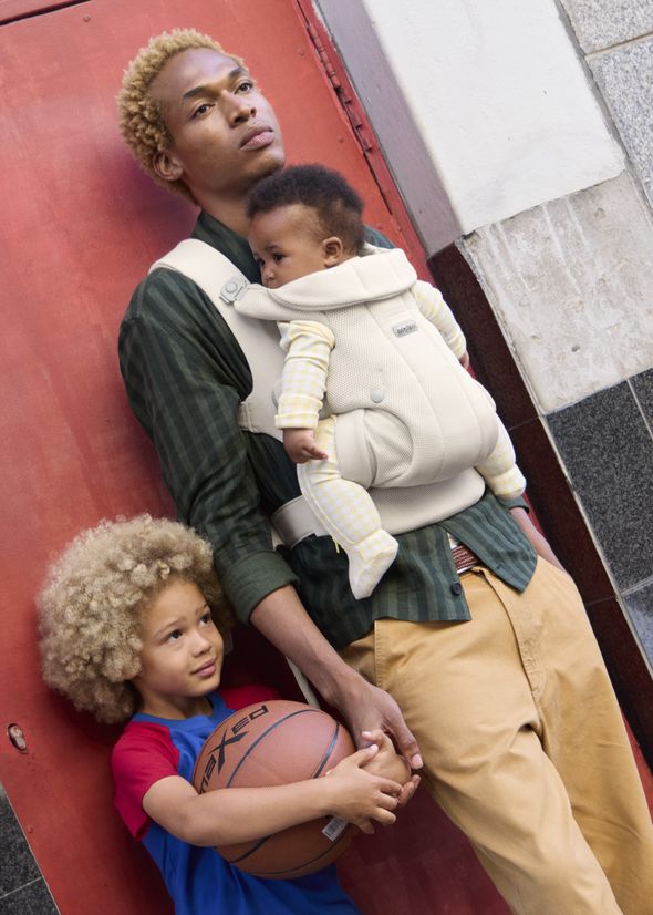 A person with blond hair leans against a red wall, carrying a baby in a front carrier, while an older child holds a basketball beside them.