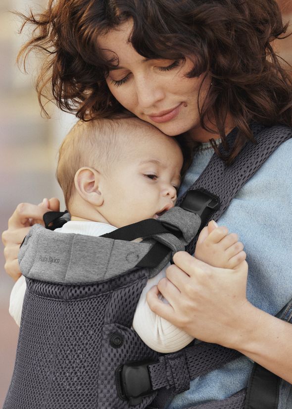 A mother lovingly holds her baby in a gray carrier. The baby rests comfortably against her chest, and they both appear content and serene.