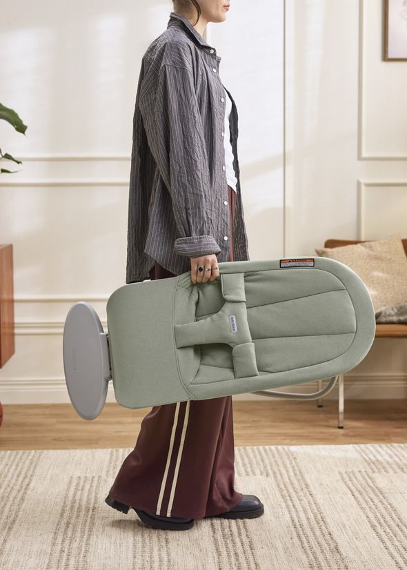 Person carrying a folded sage-green baby chair in a modern room with wooden flooring and a cozy armchair in the background.