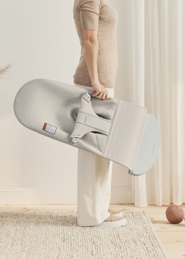 Person holding a gray baby bouncer by BabyBjörn, standing in a softly lit room with light-colored decor.