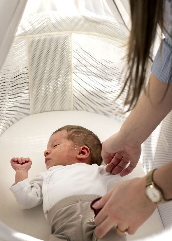 A baby wearing a white shirt and beige pants lies in a white bassinet as an adult's hands adjust the clothing.