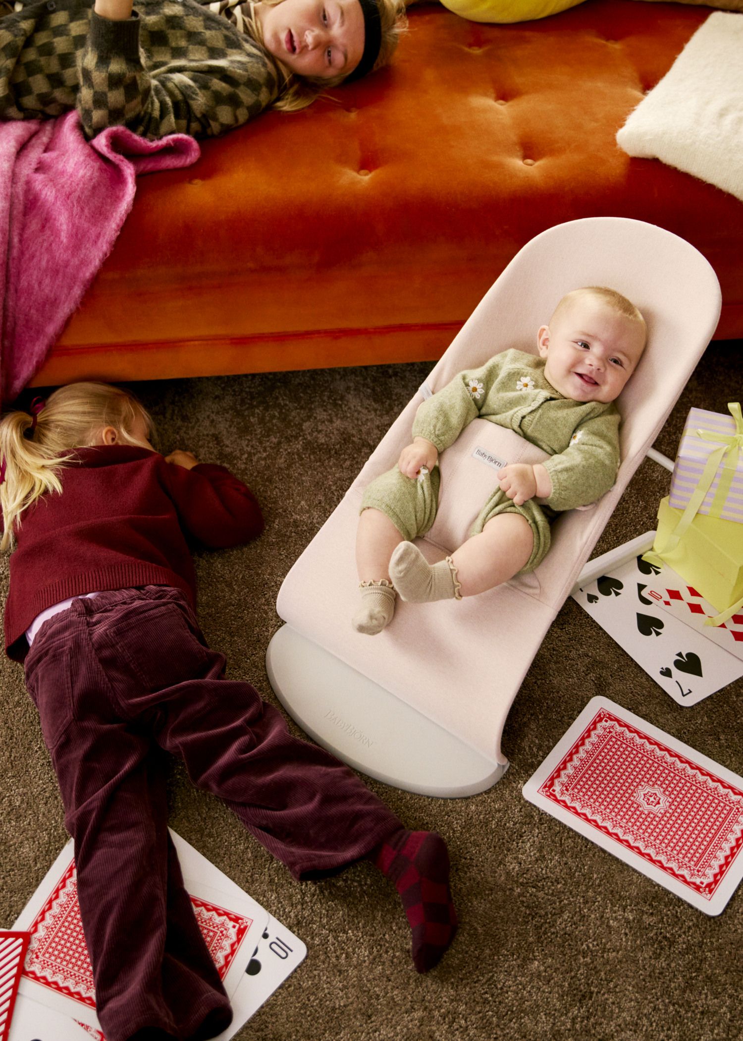 A baby in a green outfit smiles in a bouncer, surrounded by playing cards. Two children lie on the floor nearby.
