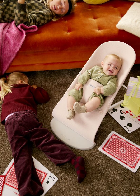 A baby in a green outfit smiles in a bouncer, surrounded by playing cards. Two children lie on the floor nearby.