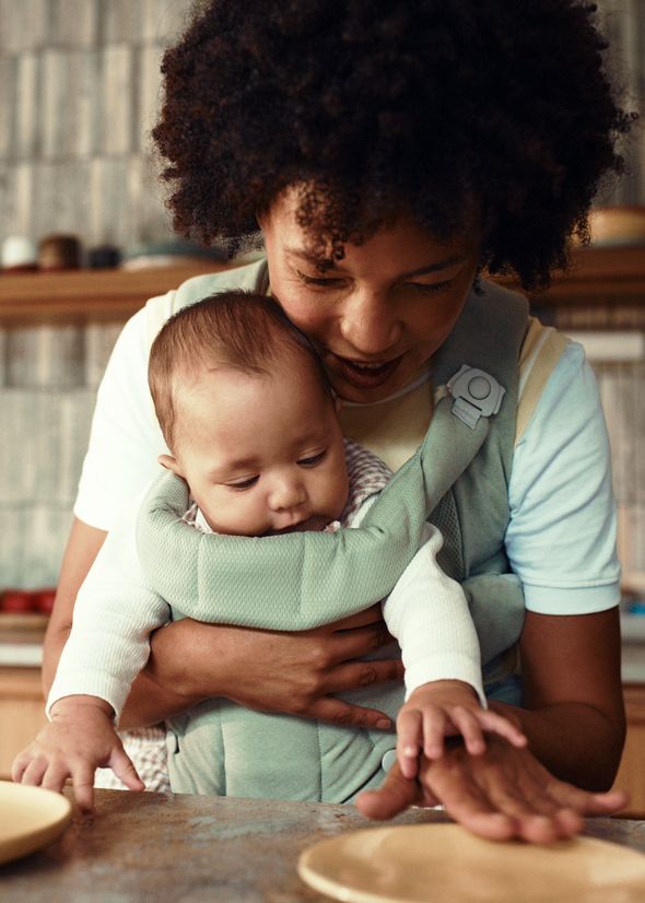 A person with curly hair holds a baby in a carrier, guiding the baby's hand on a kitchen counter, both focused on an activity.