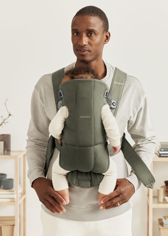 A man stands indoors wearing a gray sweatshirt, holding a baby in a green front carrier. Shelves with decorative items are visible in the background.
