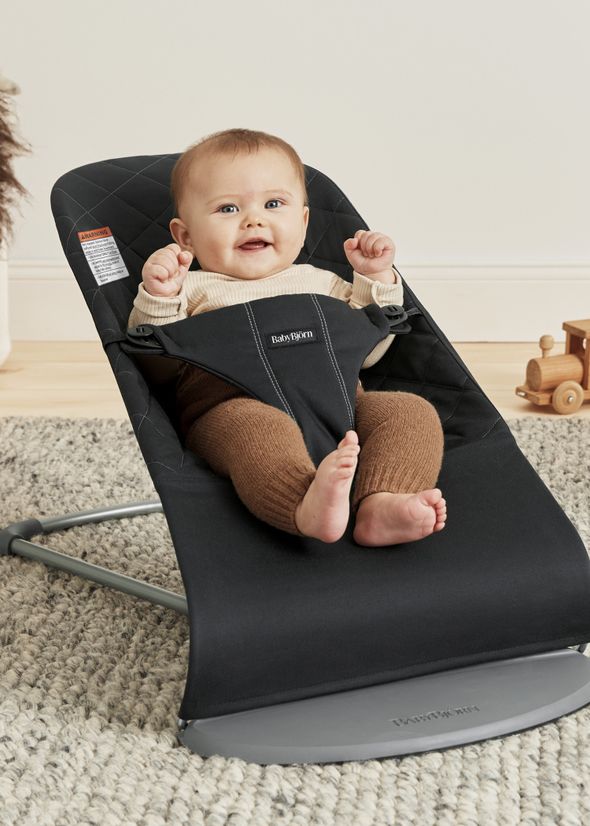 A happy baby sits in a black baby bouncer on a gray carpet, surrounded by a wooden toy train and a soft, neutral-toned background.